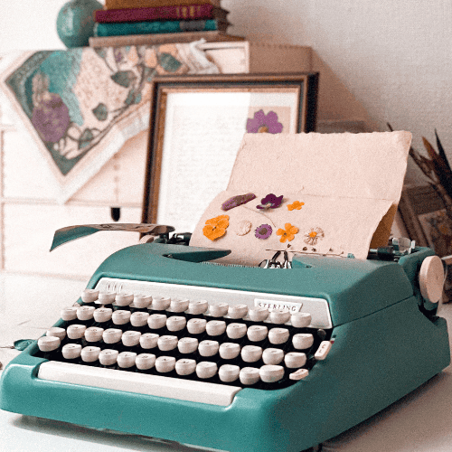 A vintage green typewriter holds a paper with pressed flowers. Behind it, framed artwork and colorful books are neatly arranged.