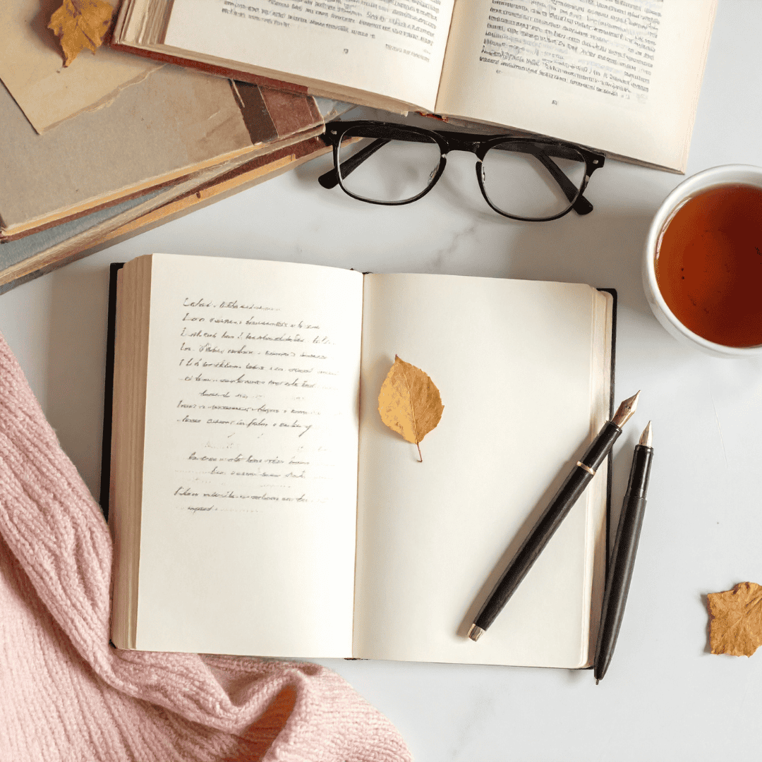 Open notebook with handwritten notes, fountain pens, glasses, and tea on a table with dried leaves and a pink scarf. Cozy autumn atmosphere.