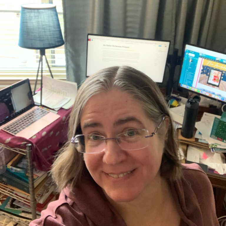 A person smiles near a workstation with multiple monitors, a laptop, documents, and a lamp in a home office setting.