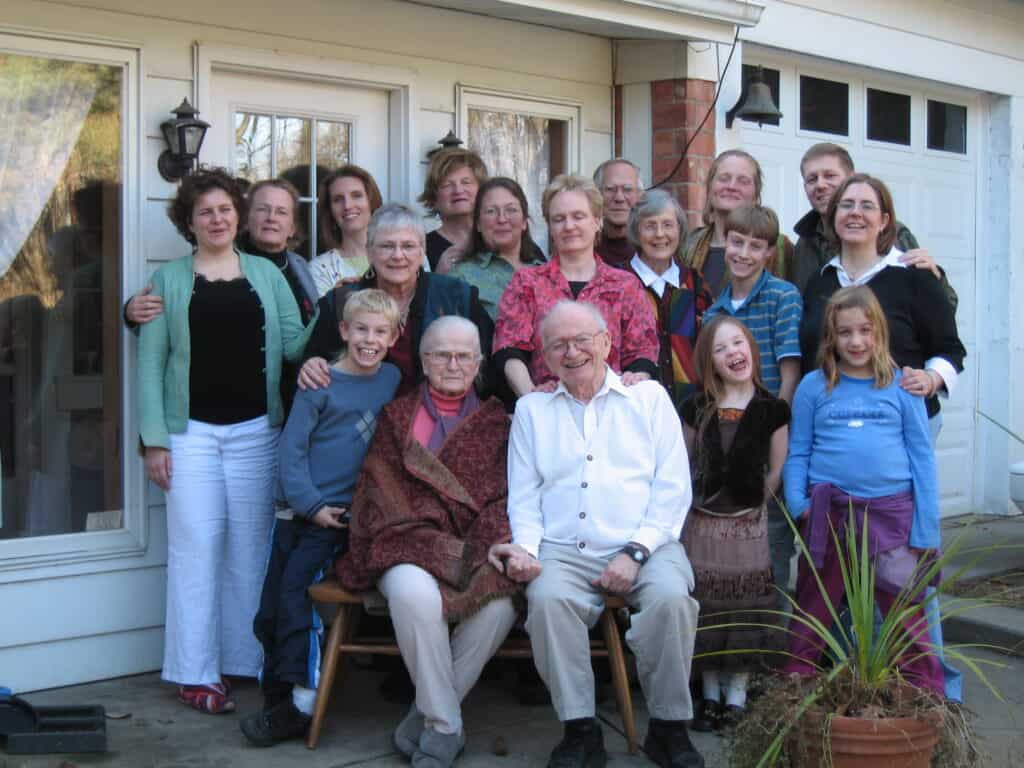 A group of people, including children and seated elders, gather outside a house with white siding and glass doors, posing for a photo.